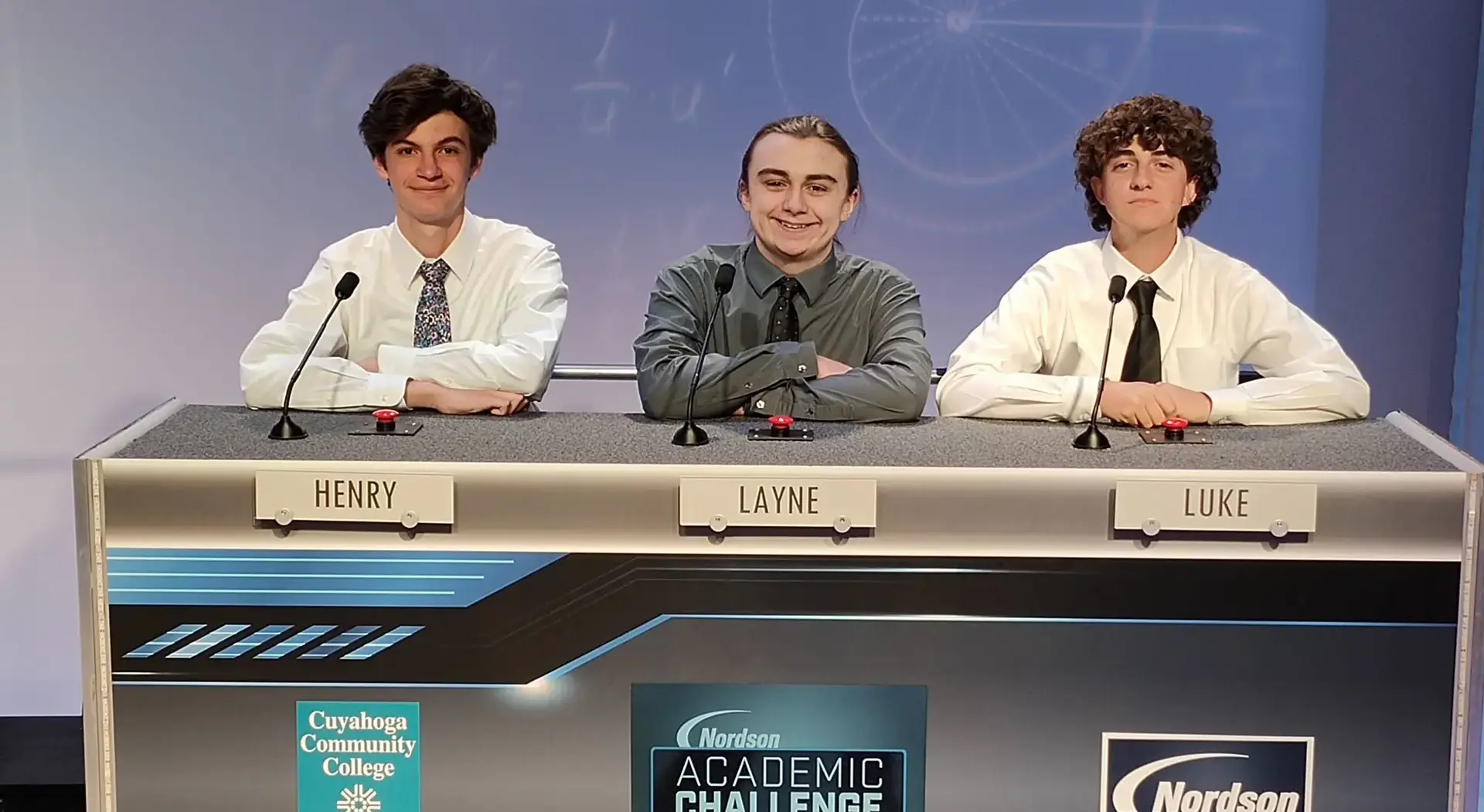 Three students seated behind microphones and buzzers at a table during an academic challenge competition