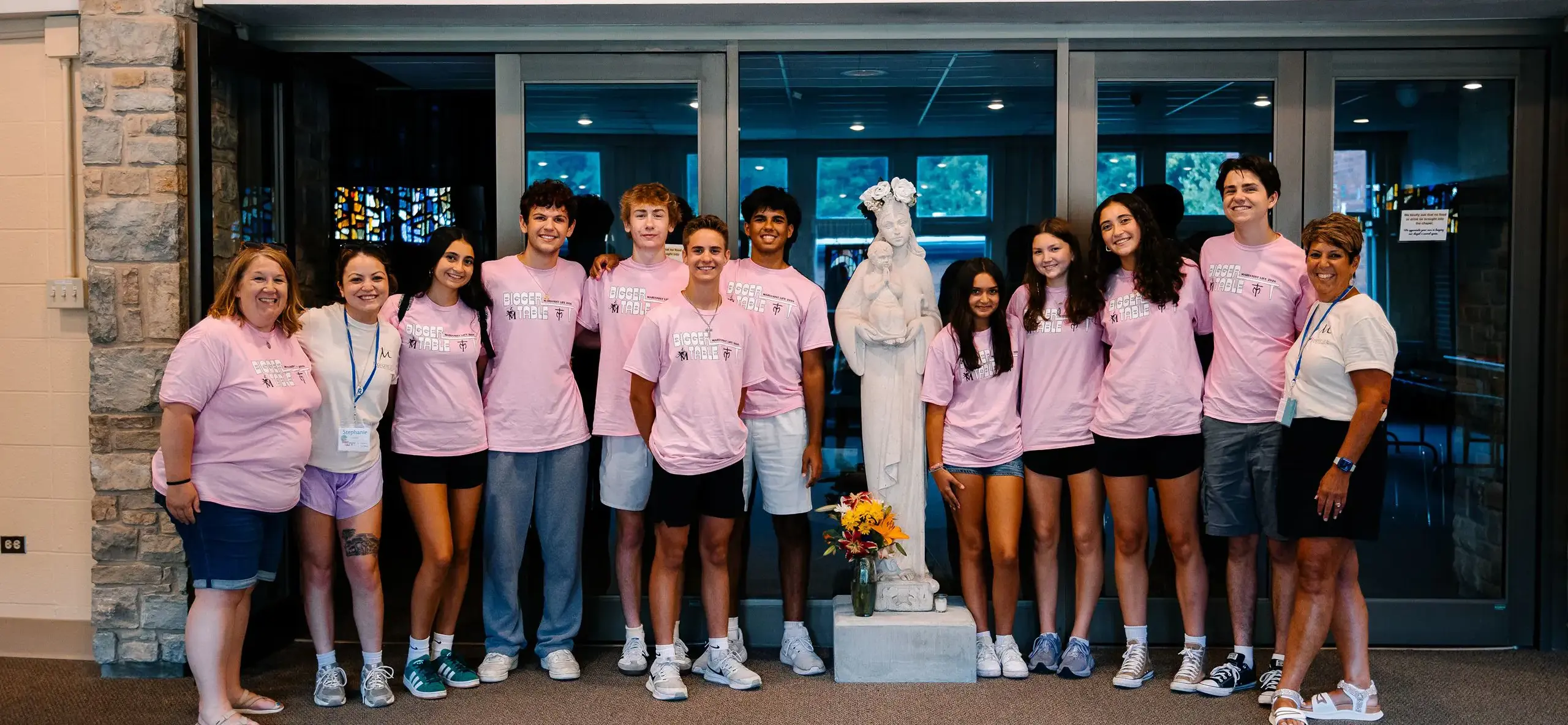 STVM students and teachers wearing pink t-shirts reading 'bigger table' stand for a photo around a statue of St. Mary.