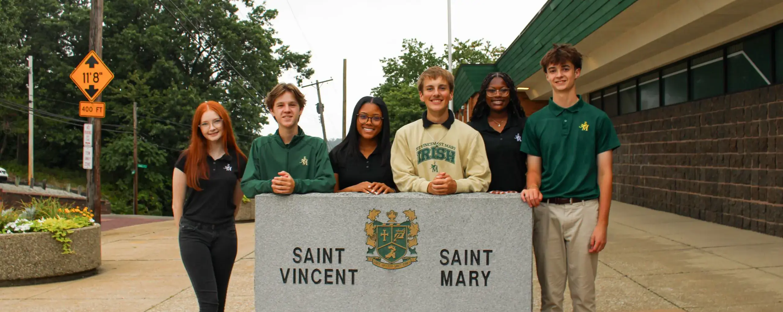 STVM students standing outside school behind a granite sign with the school's name inscribed on it.